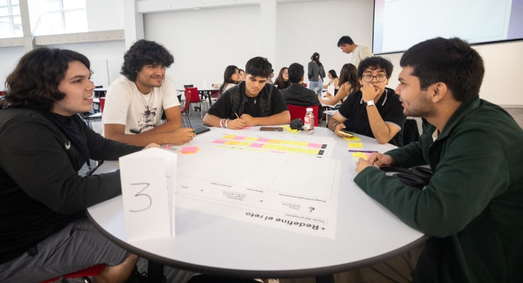 Estudiantes reunidos alrededor de una mesa redonda trabajando en equipo con una hoja de trabajo y notas adhesivas durante un taller universitario