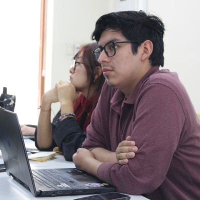 Dos estudiantes observan atentamente una presentación o clase frente a una laptop en un aula universitaria.