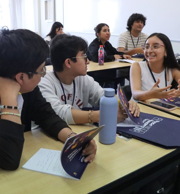 Grupo de estudiantes conversa en un aula mientras revisan materiales y folletos sobre la mesa durante una actividad académica.