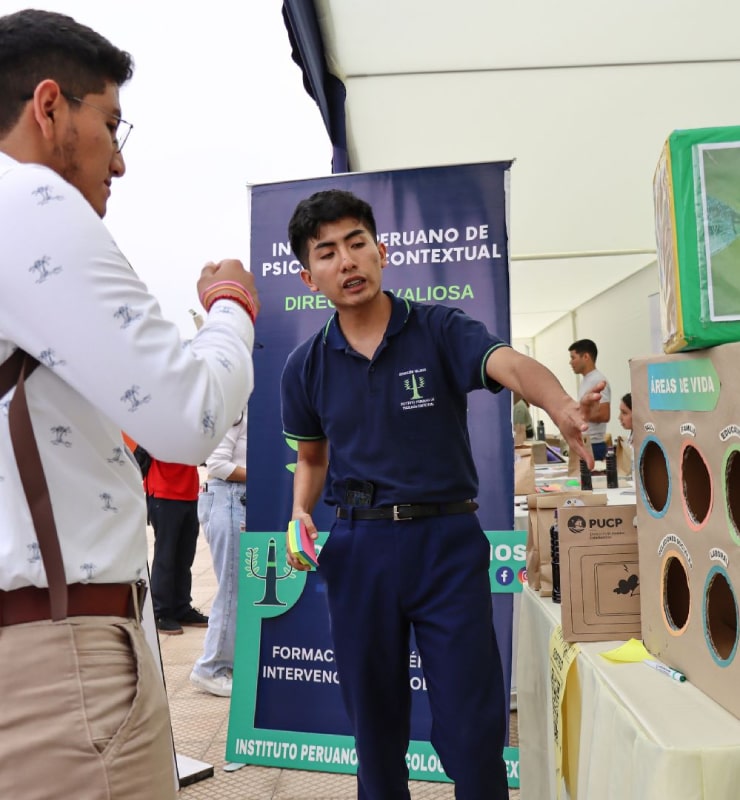 Estudiante explica un proyecto en un stand con materiales informativos y paneles del Instituto Peruano de Psicología Contextual durante una feria universitaria.