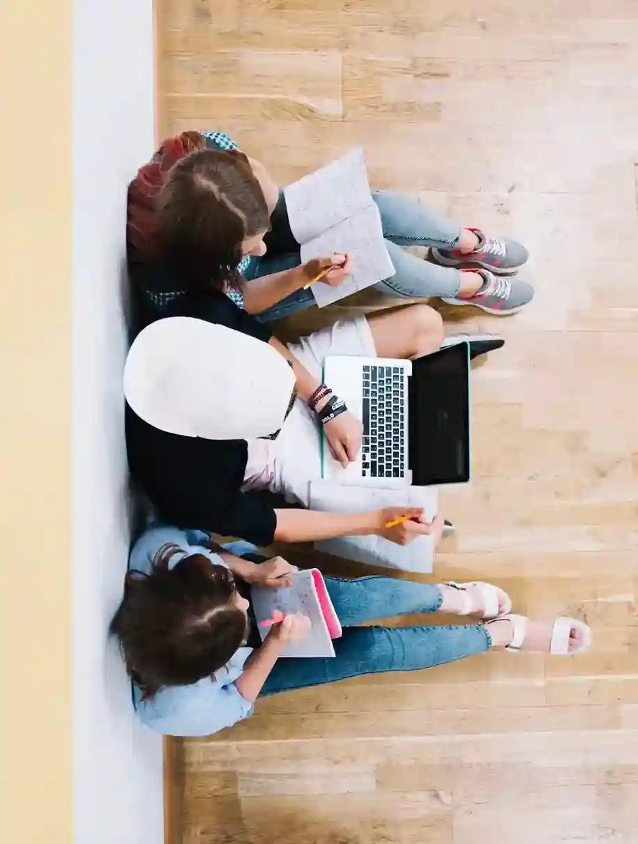 Tres estudiantes sentados en el suelo trabajando en grupo con una laptop y cuadernos, representando participación y trabajo colaborativo