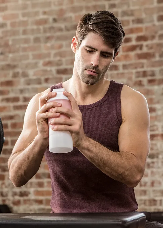 Man in sleeveless shirt preparing a protein shake at the gym, representing wellness and post-workout recovery supported by Vitamin C injections. 