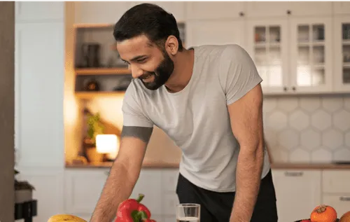 Smiling man using laptop in kitchen with fresh fruits and vegetables, representing healthy lifestyle supported by MIC lipotropic vitamin injections.