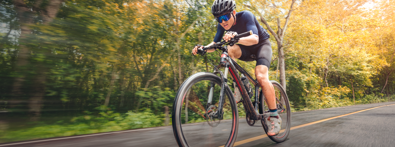Athletic man cycling on a road through a forest, wearing a helmet and sunglasses, focused on speed and performance.