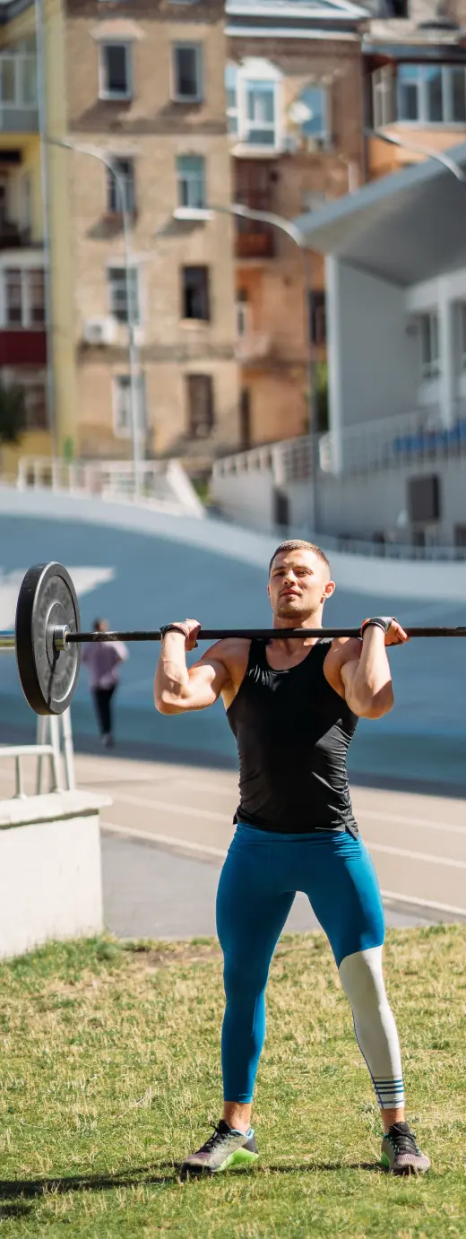 Athletic man lifting a barbell outdoors on a sunny day, wearing a black tank top and blue leggings, with buildings in the background.