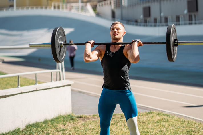 Athletic man lifting a barbell outdoors on a sunny day, wearing a black tank top and blue leggings, with buildings in the background.
