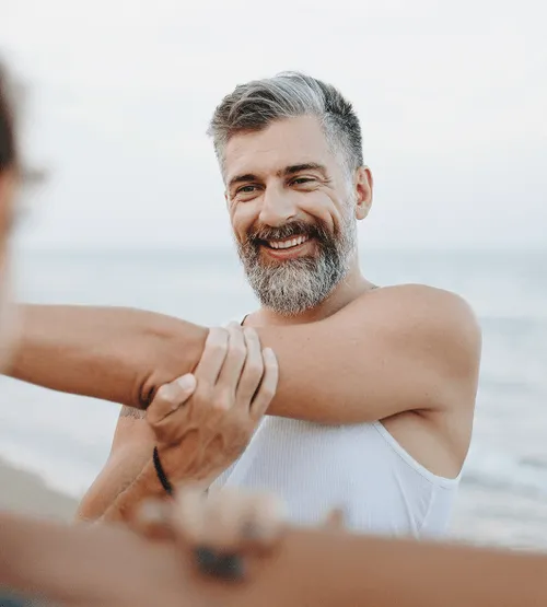 Smiling middle-aged man stretching by the beach, representing improved flexibility and vitality with Sermorelin therapy