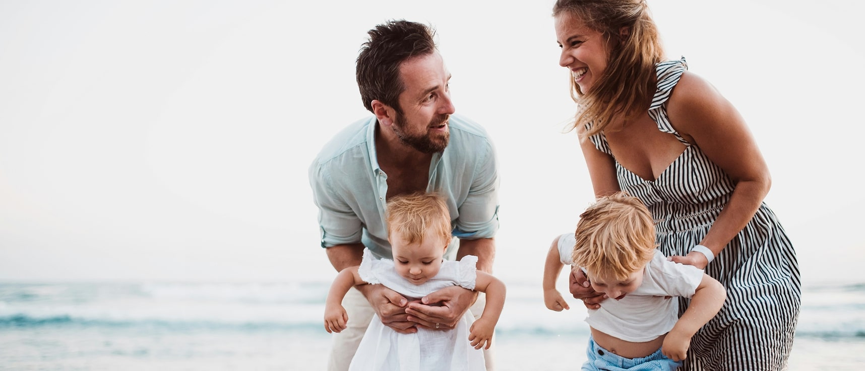 A smiling couple playing with two small children on the beach near the shoreline.