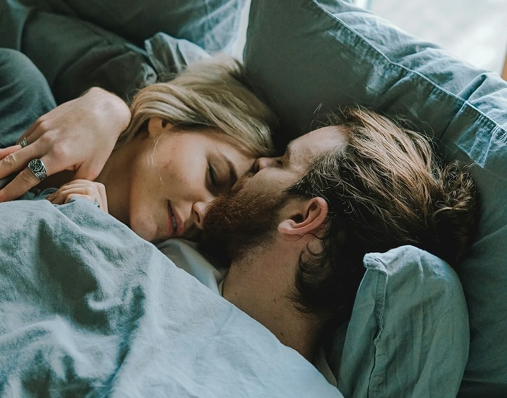 Young couple laying in bed underneath blue-gray sheets early in the morning.