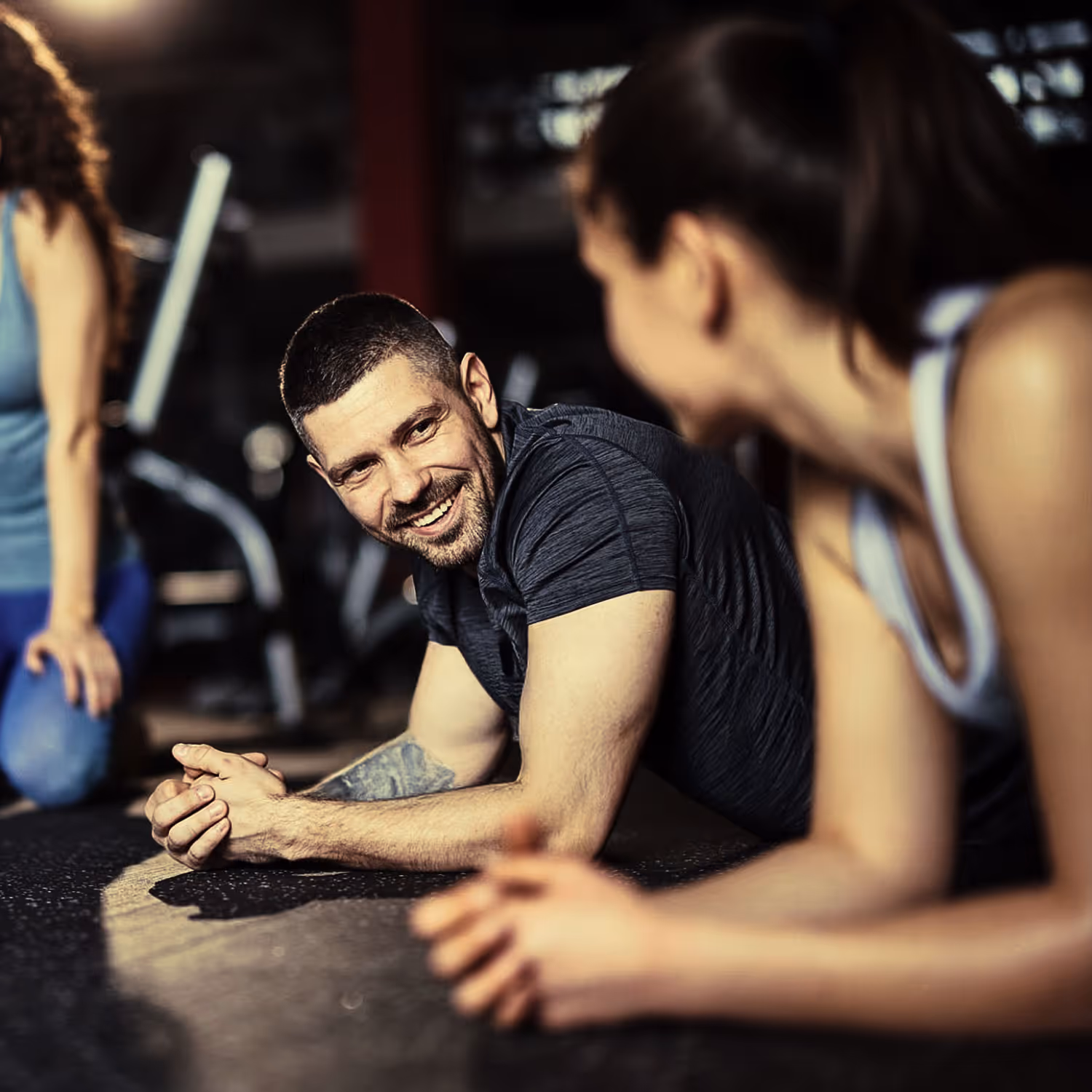 Two people in workout clothes doing planks on a gym floor, smiling and talking.