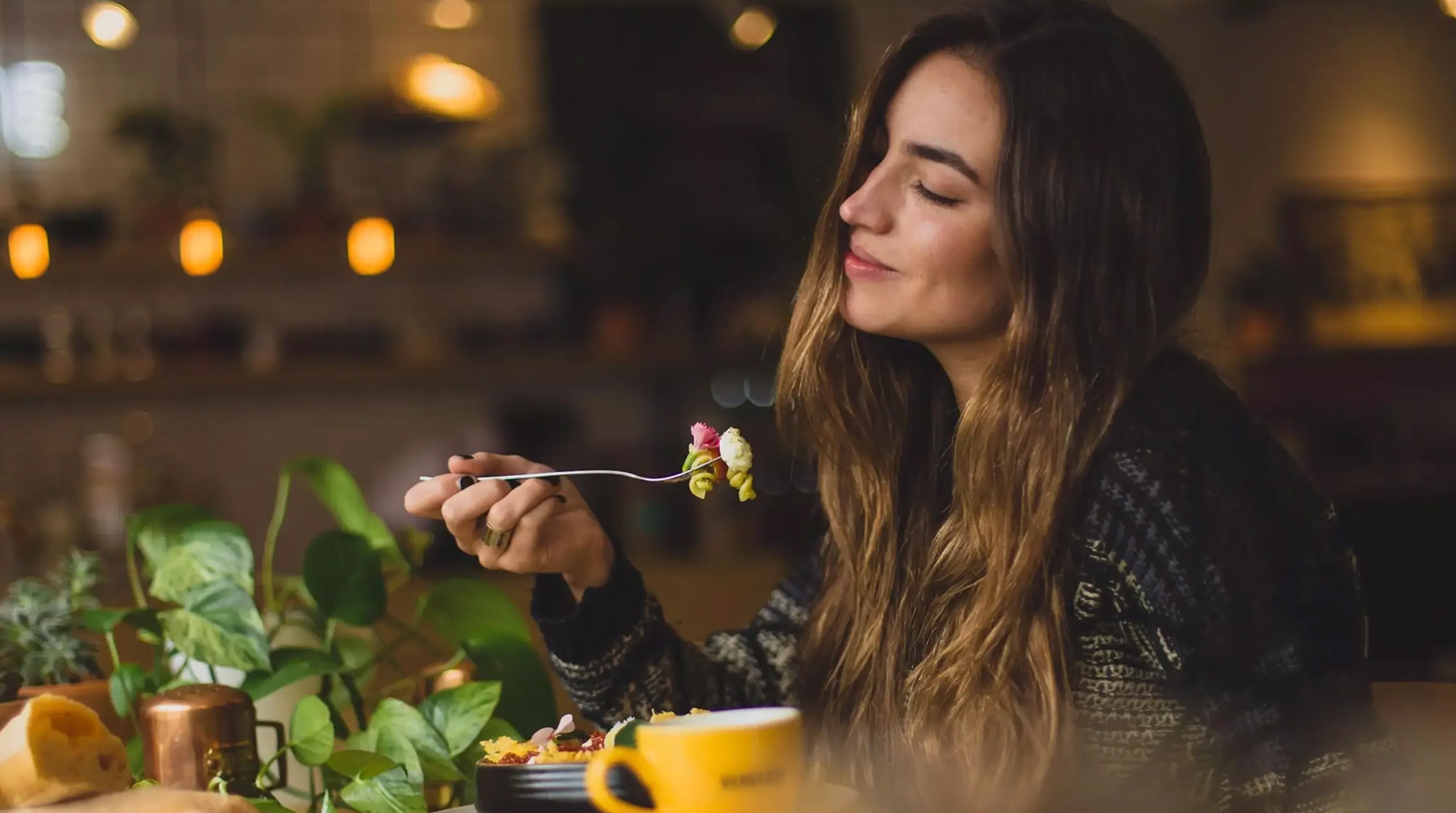 Dr. Schär Gutsense: Frau mit langen braunen Haaren genießt genussvoll eine Gabel mit Essen in einem gemütlich beleuchteten Raum.