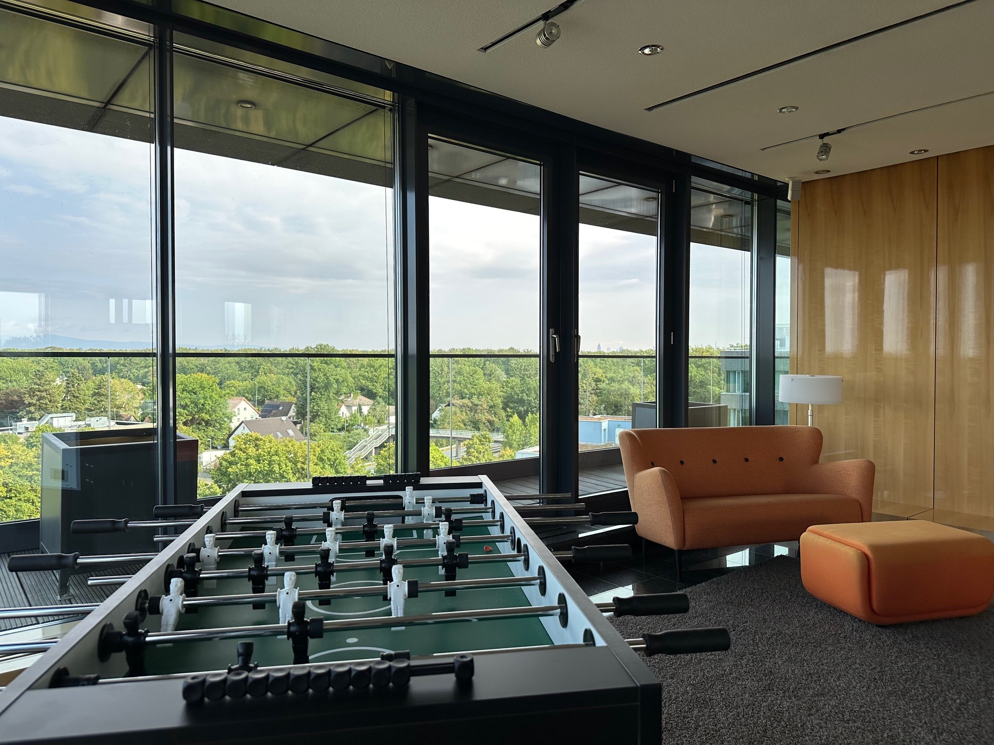 Innenraum mit einem Kicker-Tisch neben einem orangefarbenen Sofa und einem Hocker vor großen Fenstern mit Blick auf Bäume.