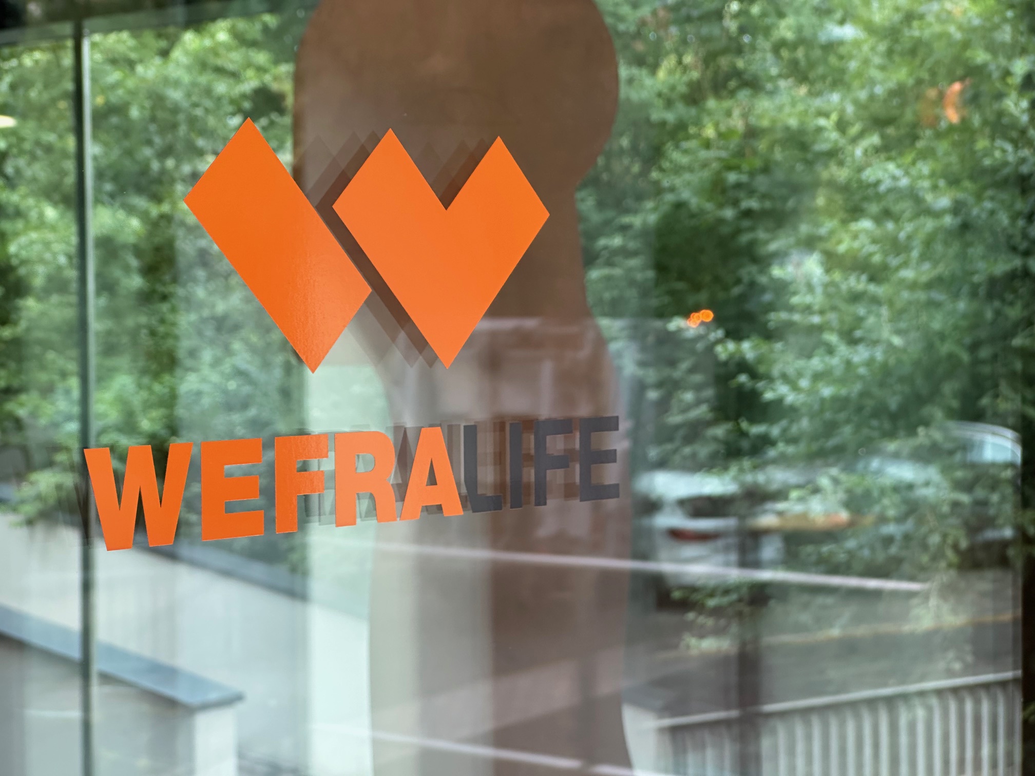 Fensterscheibe mit orangefarbenem WefraLife-Logo und grünem Wald im Hintergrund.