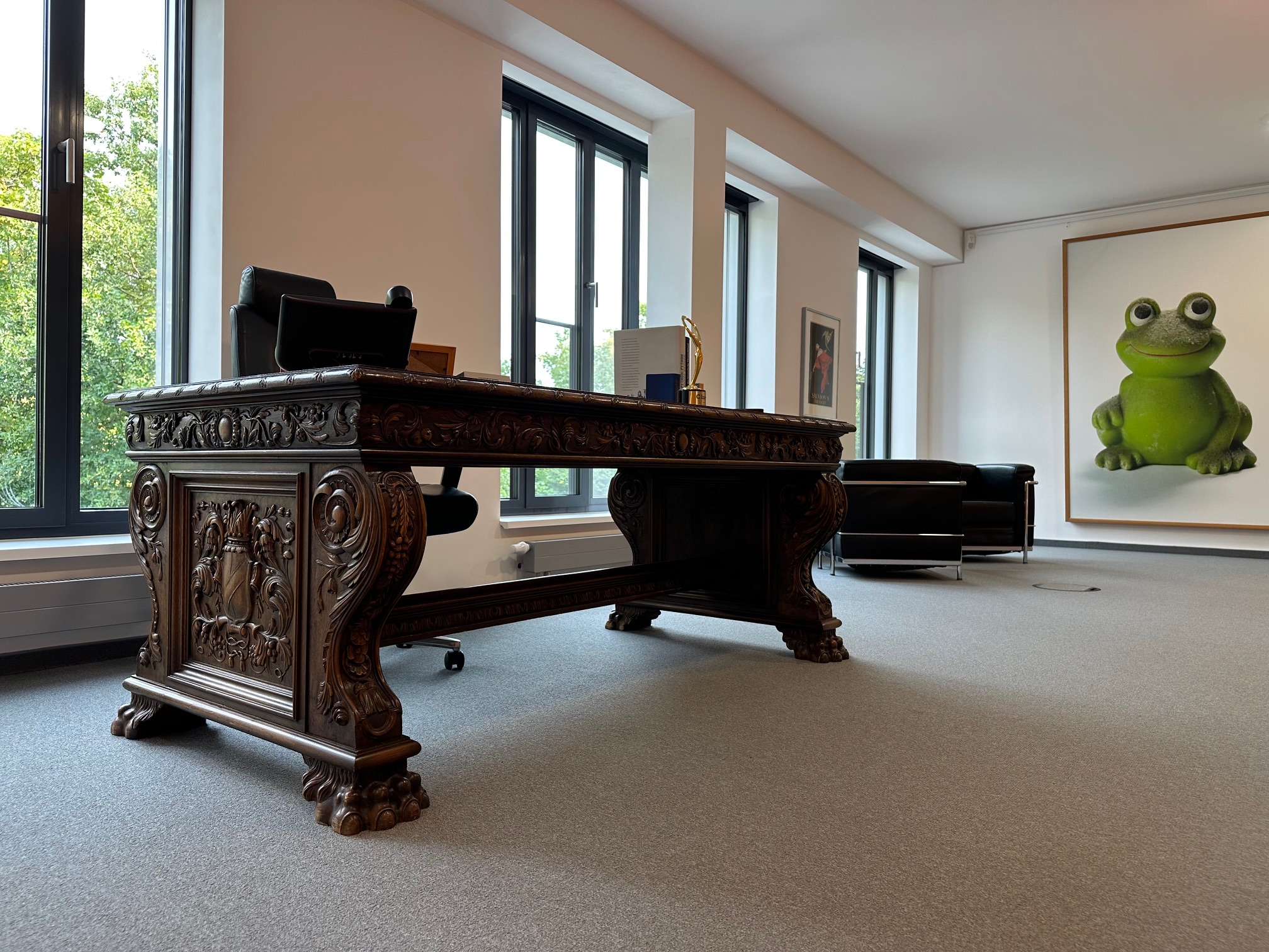 Im Büro mit einem großen, kunstvoll geschnitzten Holztisch, schwarzem Bürostuhl, zwei Ledersesseln und einem großen Bild einer grünen Froschfigur an der Wand.