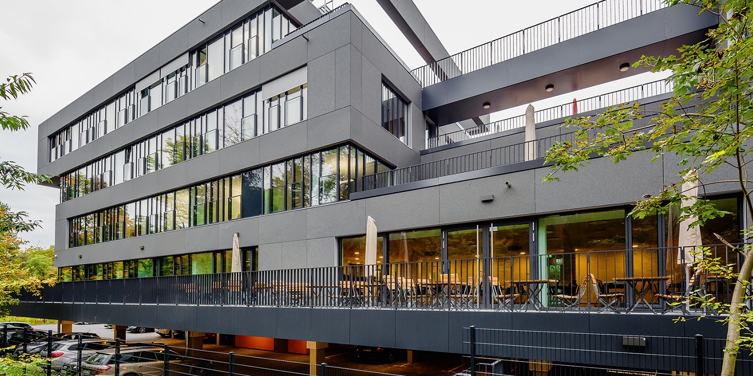 Das WEFRA Haus mit Neubau: Modernes graues Bürogebäude mit großen Fensterfronten und einer überdachten Terrasse mit Tischen und Stühlen.