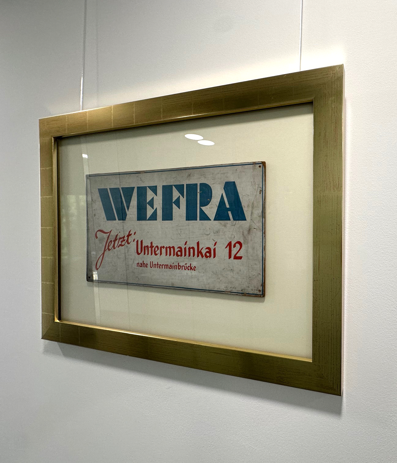 Gerahmtes Schild mit der Aufschrift 'WEFRA Jetzt: Untermainkai 12 nahe Untermainbrücke' an einer weißen Wand.