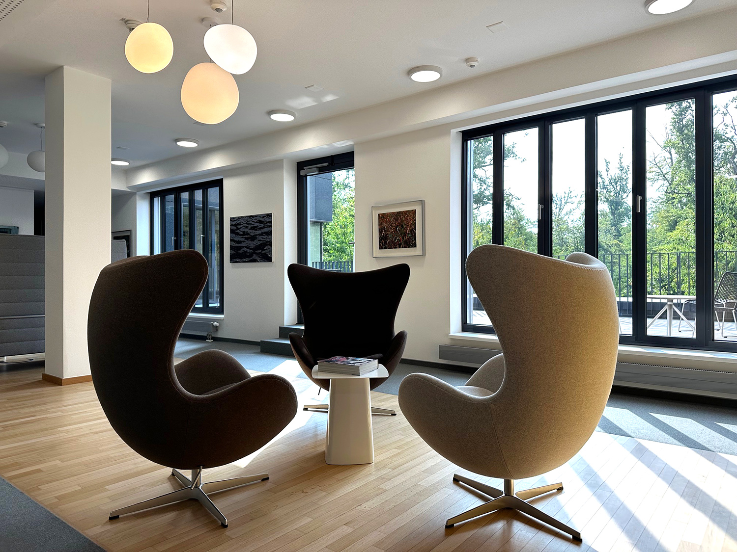 Drei moderne Eierstuhl-Sessel um kleinen weißen Tisch in einem hellen Raum mit großen Fenstern und Holzfußboden.