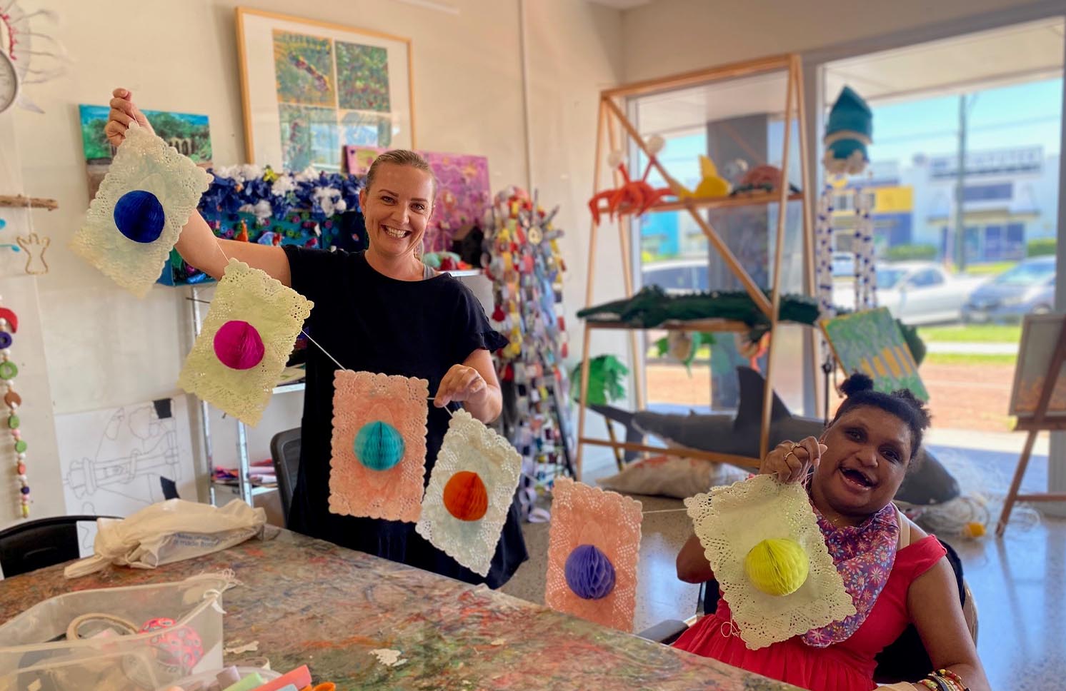 Two smiling women in an art studio, holding up handmade decorations made of lace and colored yarn balls.