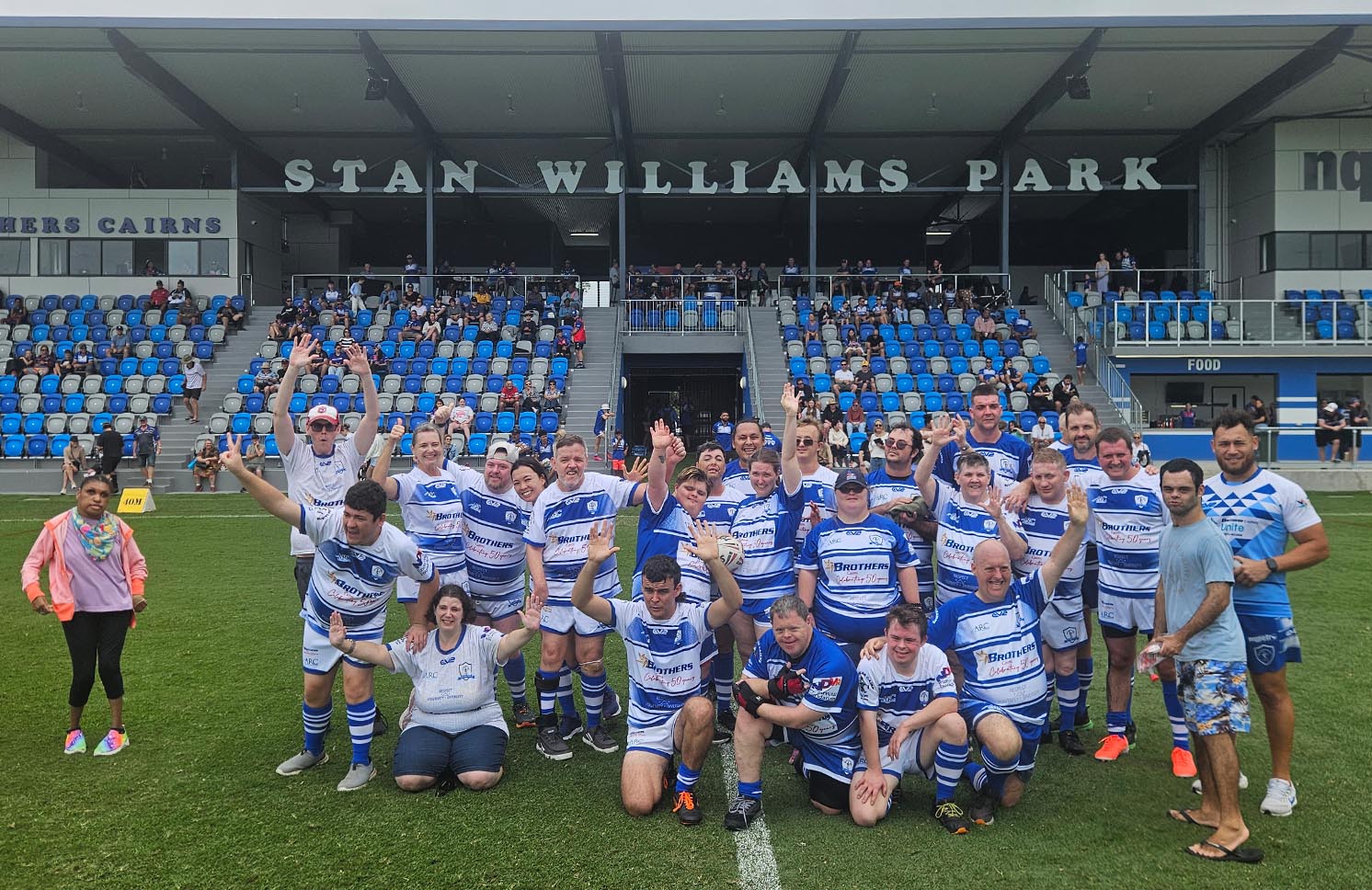 A group of people in blue and white rugby uniforms with upraised arms, celebrating on a field under the Stan Williams Park grandstand.