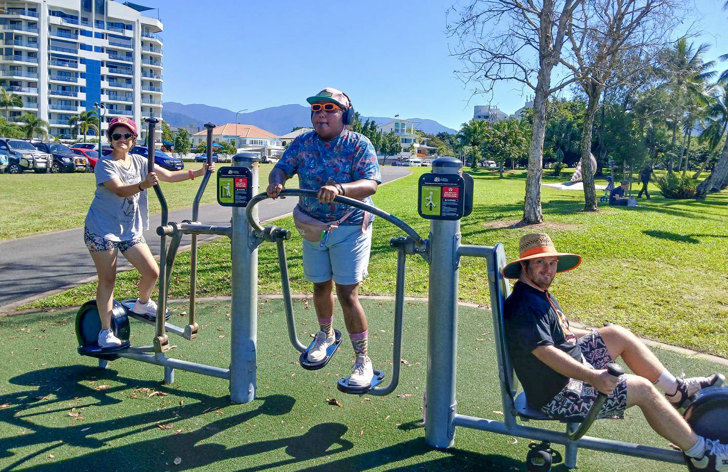 Three people exercising on outdoor fitness equipment in a sunny park with city buildings in the background.
