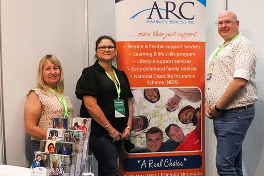 Three people standing next to an ARC Disability Services banner at a conference booth.