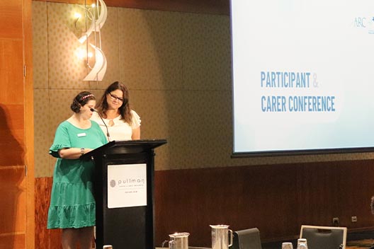 Two women speaking at a podium in front of a screen displaying Participant & Carer Conference.