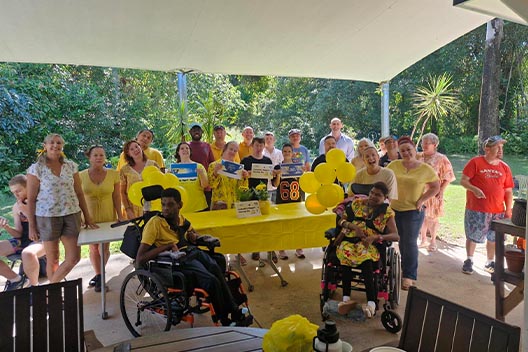 A large group of people wearing yellow for the Biggest Morning Tea, gathered under a patio awning with yellow balloons.