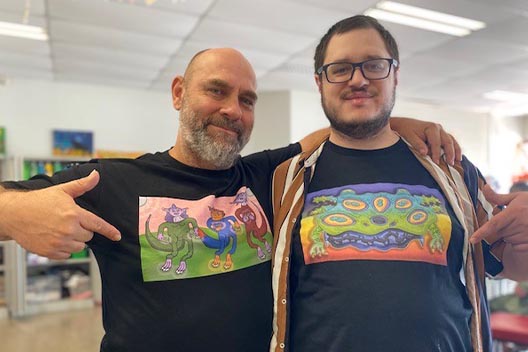 Two men smiling, pointing to colorful, custom-designed t-shirts featuring artwork.