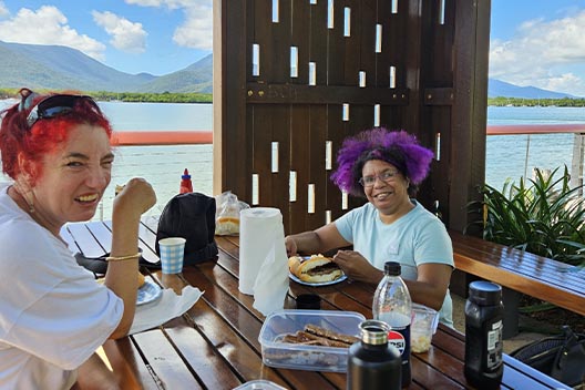 Two smiling women sitting at an outdoor picnic table with a harbor and mountains in the background.