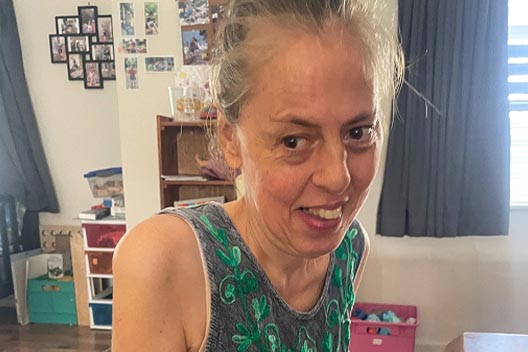 A close-up portrait of a smiling woman indoors in a grey tank top with green embroidery.
