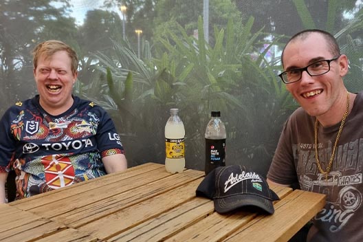 Two men sitting at a wooden table outdoors, smiling. One is wearing a rugby jersey and the other glasses and a gold chain.