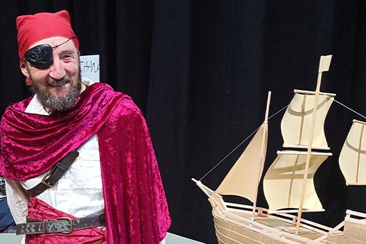 A man dressed as a pirate with an eyepatch and red bandana smiling next to a model wooden sailboat.