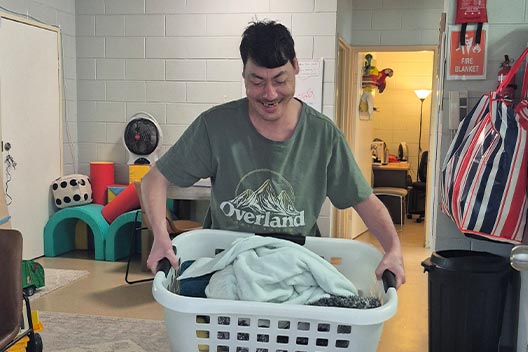A smiling man in a green shirt carrying a white laundry basket full of clothes indoors.