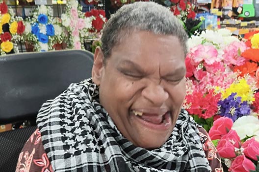A woman with a scarf smiling enthusiastically, seated in a wheelchair near a colorful display of flowers.