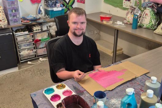 A man smiling while seated at a table in an art studio, painting a patch of pink on a piece of cardboard.