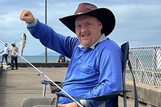 A man in a hat sitting on a pier, smiling while proudly holding up a small fish he just caught.