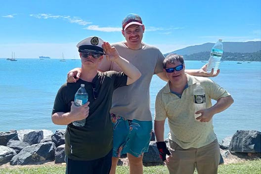 Three men standing by the water on a sunny day, holding water bottles. One is wearing a captain's hat and saluting.