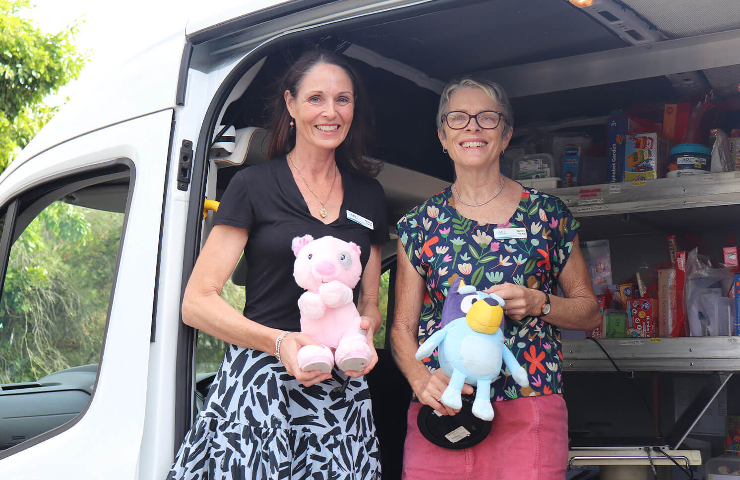 Two women in the back of a white van smiling and holding stuffed toys