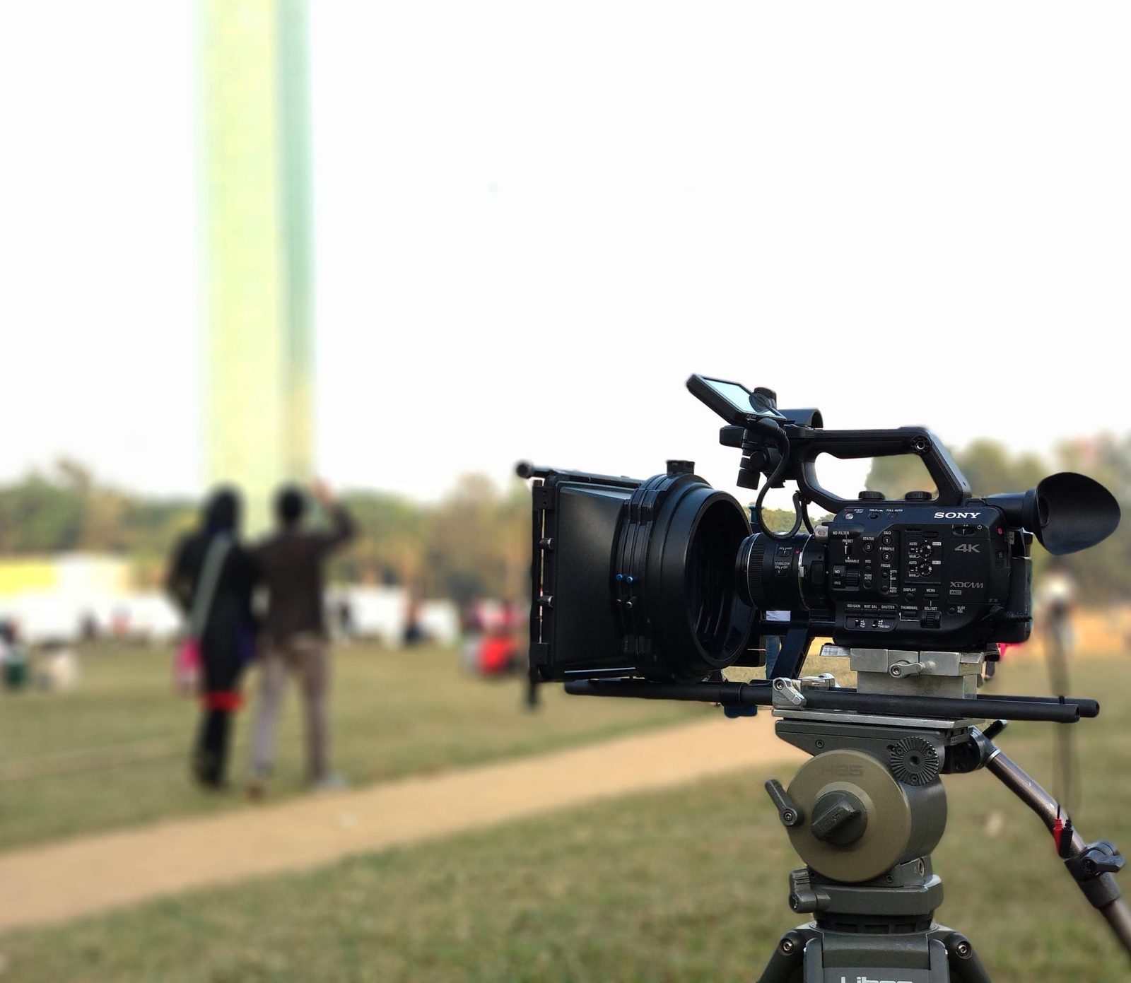 A professional 4K cinema camera with a matte box on a tripod at an outdoor shoot, crew blurred in the background