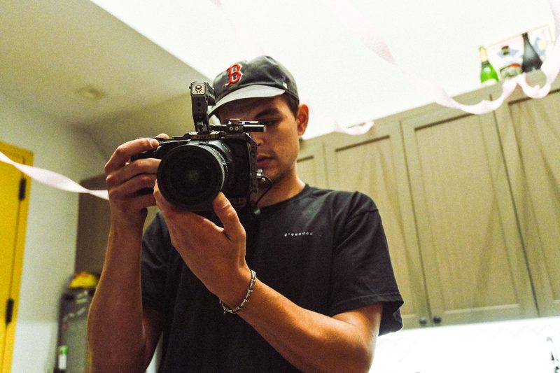 A videographer in a cap holds a rigged mirrorless camera, steadying it with both hands to film indoors.