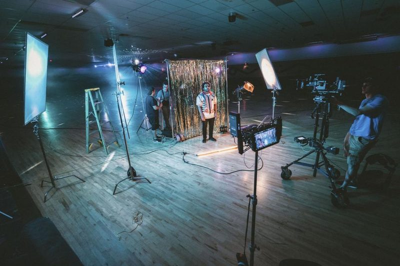 A video crew films a person on a lit studio set with softbox lights, a cinema camera on a tripod, and light stands.