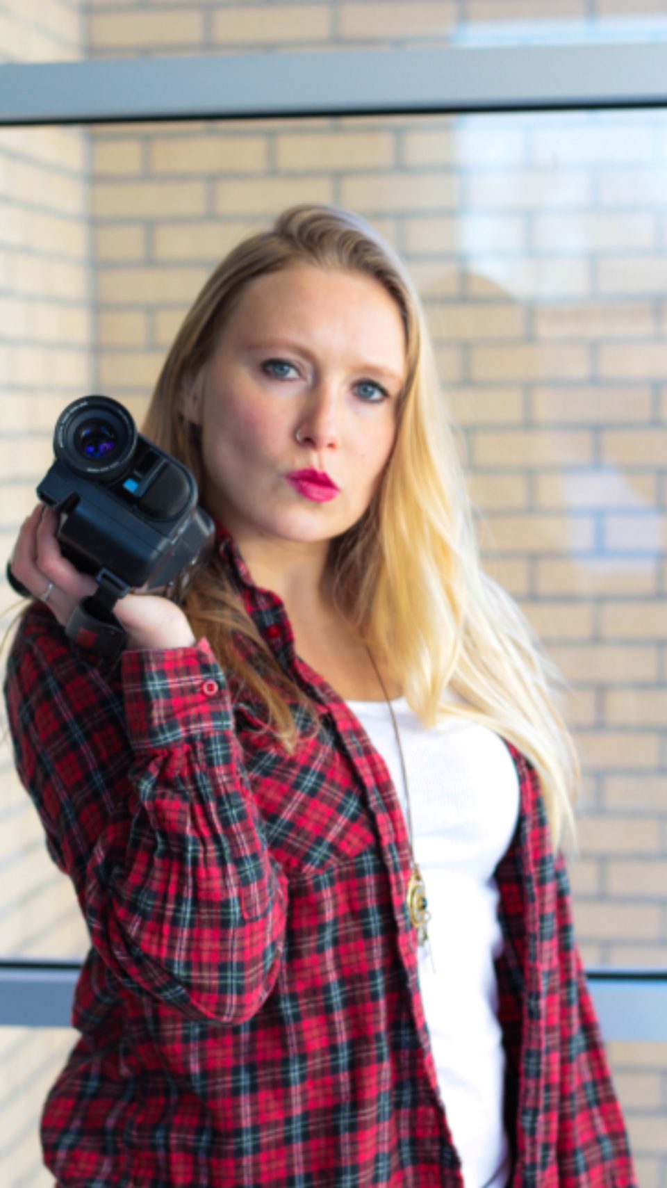 A person in a red plaid shirt holding a handheld video camera in front of a brick and glass wall