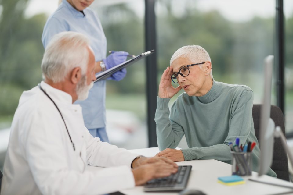 A doctor and a nurse review notes with an older patient during a consultation in a bright medical office