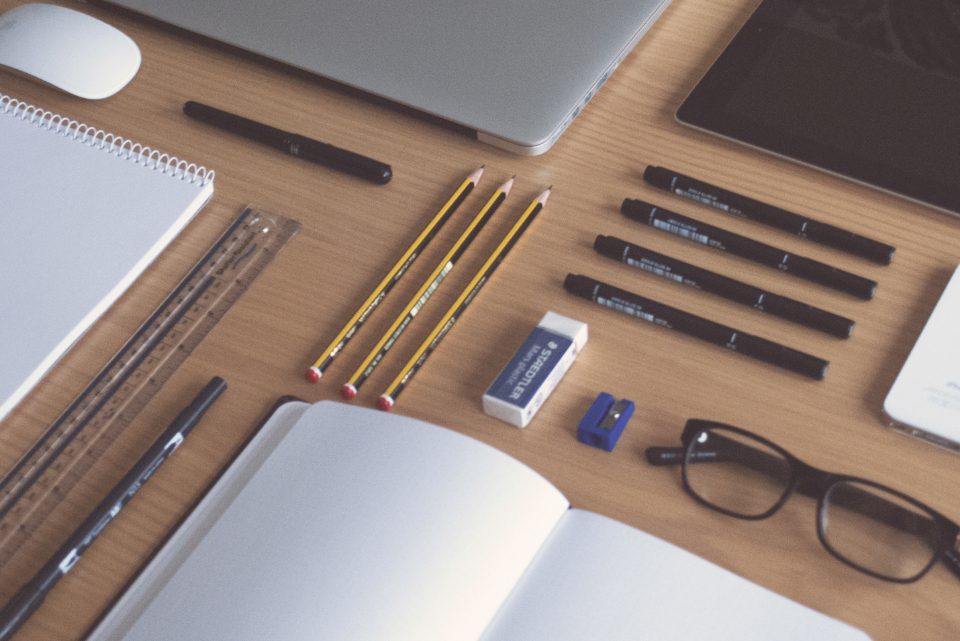 Flat-lay of a wooden desk with a laptop, notebook, pencils, pens, ruler, and glasses arranged for planning.