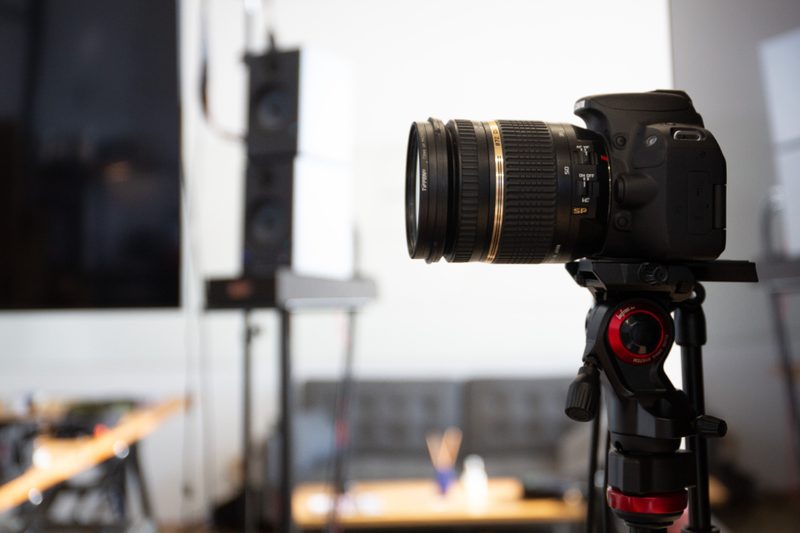 A video camera on a tripod on a lit studio set, with a softbox light glowing behind it.