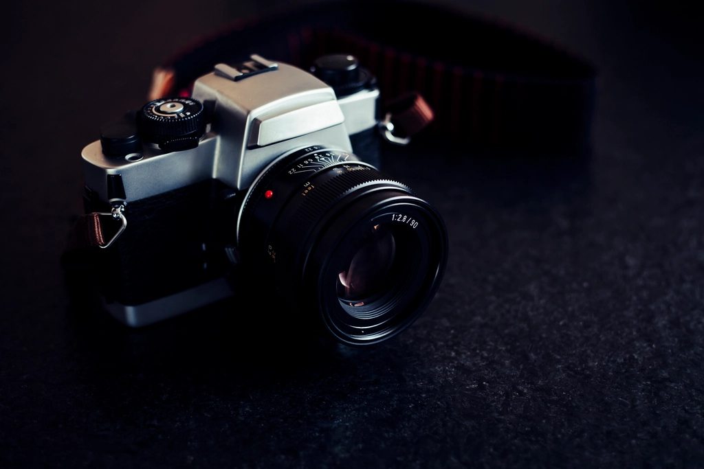 A silver and black 35mm SLR film camera with a lens, resting on a dark surface with its strap coiled behind it.