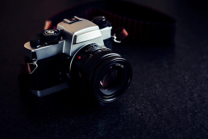 A silver and black 35mm SLR film camera with a lens, resting on a dark surface with its strap coiled behind it.