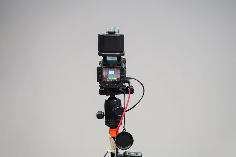 A professional video camera on a tripod with cables connected, its screen showing a live preview of the shot