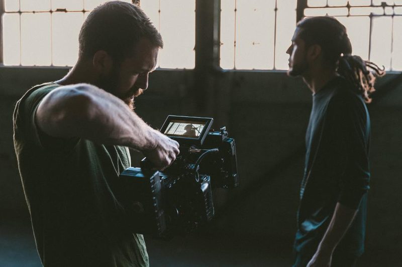 A camera operator adjusts a cinema camera with a monitor while filming a man on an indoor set.