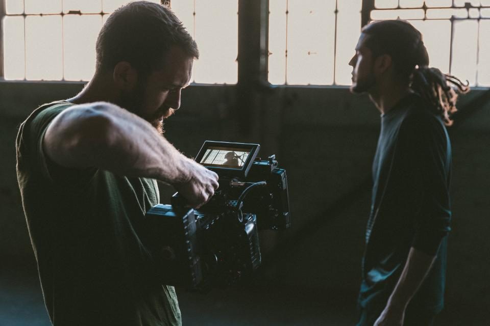 A camera operator adjusts a cinema camera with a monitor while filming a man on an indoor set.